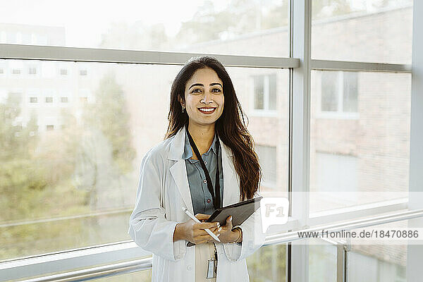 Porträt einer lächelnden Ärztin  die ein digitales Tablet und einen Stift hält und am Fenster eines Krankenhauskorridors steht
