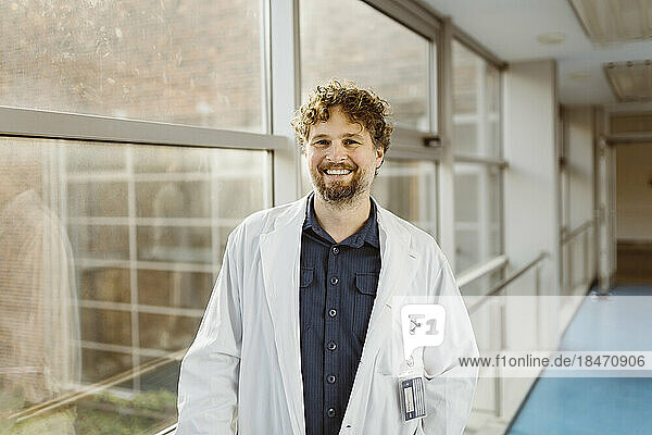 Porträt eines lächelnden reifen Arztes in einem Laborkittel  der in einem Krankenhauskorridor am Fenster steht
