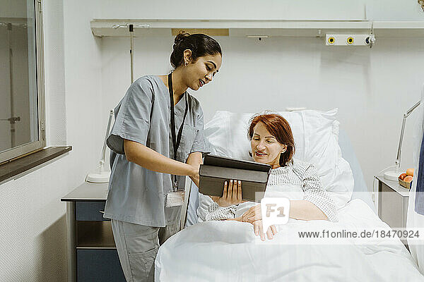Smiling nurse showing digital tablet to senior female patient lying on bed in hospital