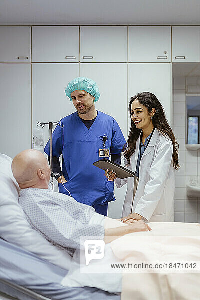 Lächelnde Ärztin  die einen älteren männlichen Patienten tröstet  der von einem Kollegen im Krankenhaus auf dem Bett liegt