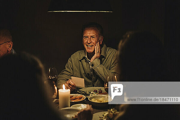 Lächelnder älterer Mann genießt ein Abendessen bei Kerzenlicht am beleuchteten Tisch