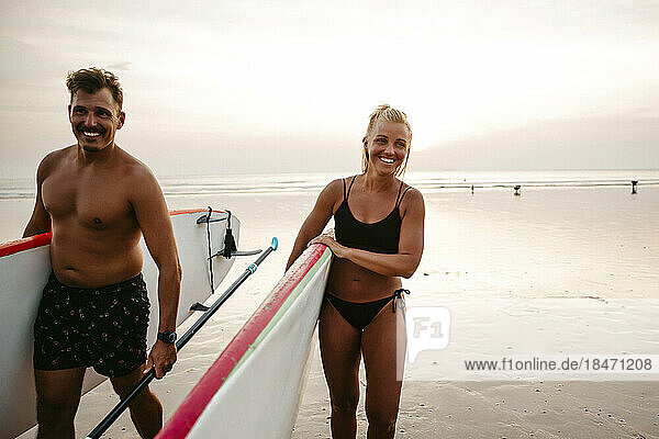 Happy couple with paddleboards walking at beach during sunset