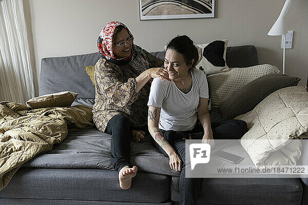 Glückliche Frau mit Querschnittslähmung wird von ihrer Mutter massiert  während sie zu Hause auf dem Sofa sitzt