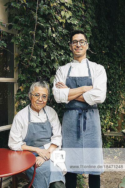 Porträt eines selbstbewussten männlichen Kochs  der neben einer älteren Kollegin vor einem Restaurant steht