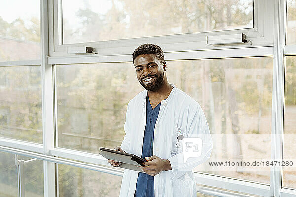 Porträt eines lächelnden jungen Arztes  der ein digitales Tablet hält und am Fenster eines Krankenhauskorridors steht