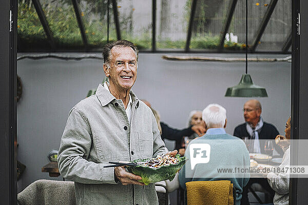 Porträt eines lächelnden älteren Mannes  der eine Salatschüssel bei einer Dinnerparty hält
