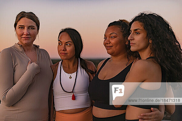 Multiracial female friends standing with arms around at sunset
