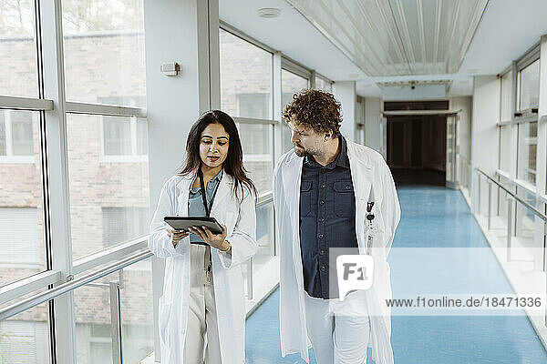 Eine Ärztin diskutiert mit einem männlichen Kollegen über ein digitales Tablet  während sie gemeinsam durch einen Krankenhausflur gehen