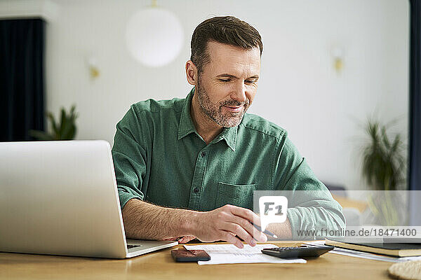 Mann sitzt zu Hause mit Laptop und Dokumenten und prüft die Finanzen seines Hauses