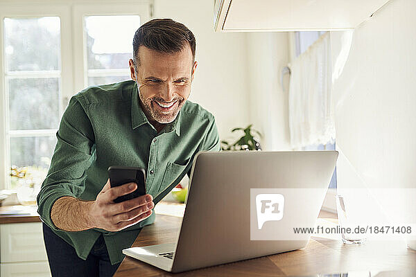 Smiling man making online payment with mobile phone and laptop in the kitchen