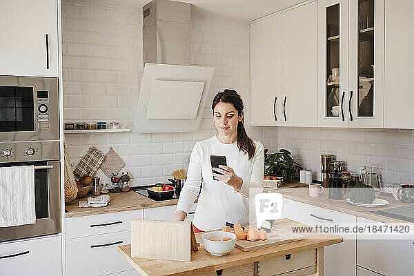 Lächelnde Frau benutzt Mobiltelefon in der heimischen Küche
