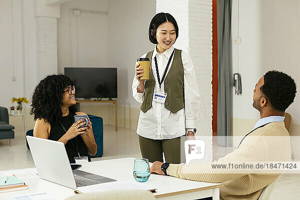 Happy woman with disposable cup talking with colleague in office