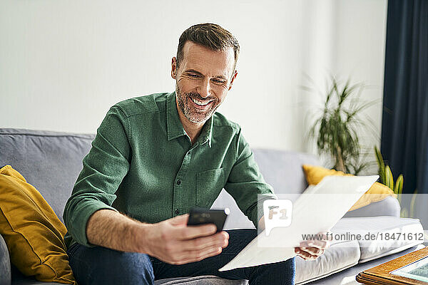 Smiling man sitting on couch paying bills with his smartphone