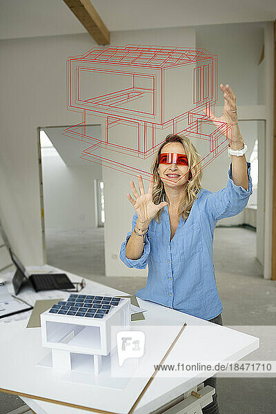Smiling mature architect wearing virtual reality simulator gesturing and examining house model