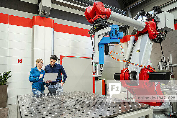 Technician with laptop standing by colleague in robot factory