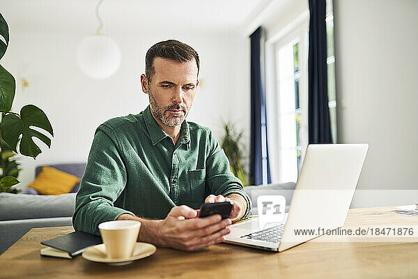 Mann benutzt Telefon  während er von zu Hause aus arbeitet und am Tisch mit Laptop sitzt