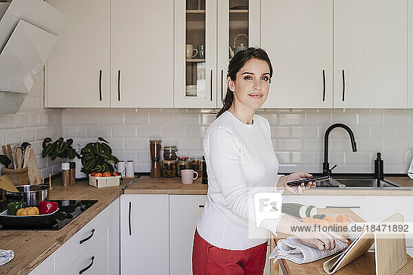 Smiling woman with tablet PC holding smart phone in kitchen at home