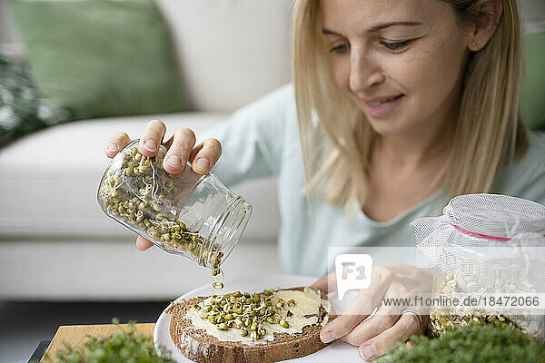 Woman pouring sprouts from glass jar on toasted bread at home