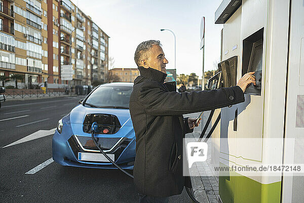 Mature businessman charging electric car at station on sunny day