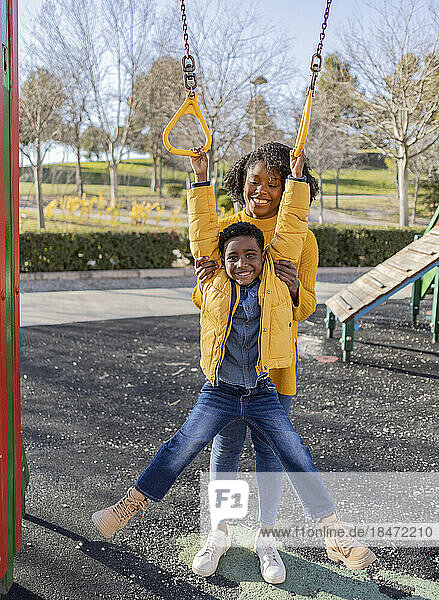 Happy mother supporting son hanging on gymnastic rings at playground
