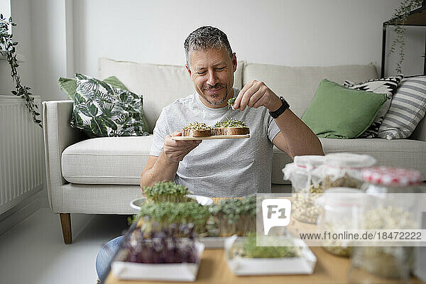 Smiling man eating sprouts sitting in front of sofa at home