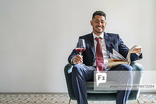 Happy businessman sitting with book and glass of red wine on armchair in front of wall