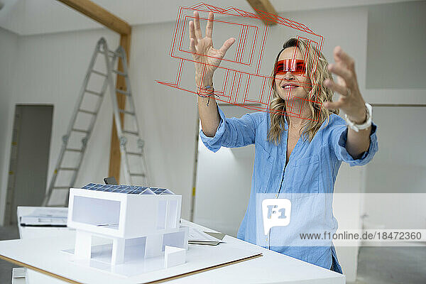 Mature architect wearing virtual reality simulator gesturing and examining house model at site