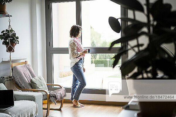 Woman with coffee cup standing near window at home