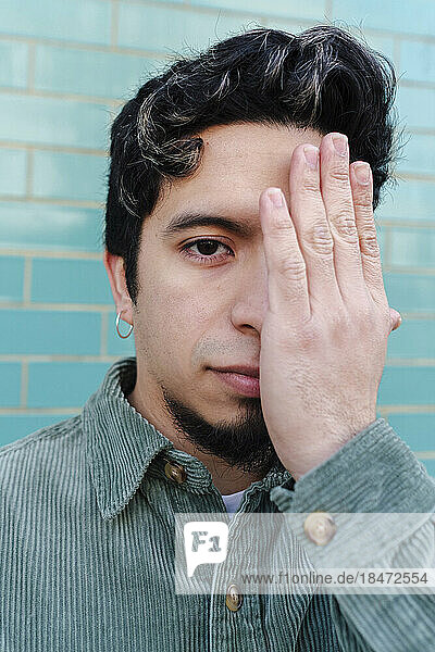 Mann bedeckt Auge mit Hand vor türkisfarbener Ziegelwand