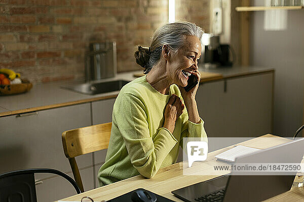 Happy freelancer talking on smart phone at desk in home