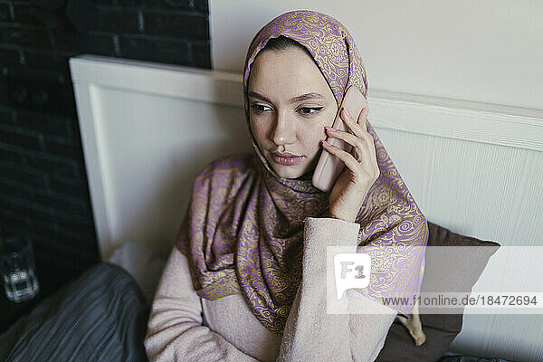 Young woman wearing hijab talking on smart phone in bed at home
