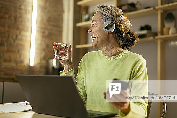 Fröhliche reife Frau mit Smartphone sitzt am Schreibtisch