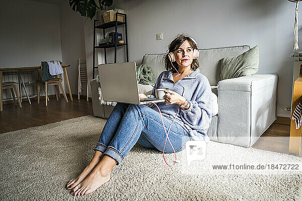 Nachdenkliche Frau sitzt mit Laptop und Kaffeetasse zu Hause