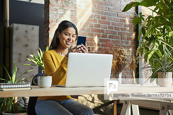 Happy businesswoman with laptop using mobile phone at desk in loft office