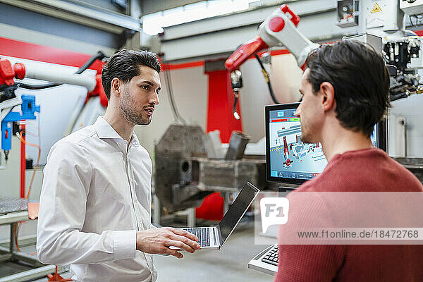 Technician holding laptop and explaining colleague at robot factory