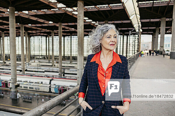 Mature businesswoman with hands in pockets walking at railroad station