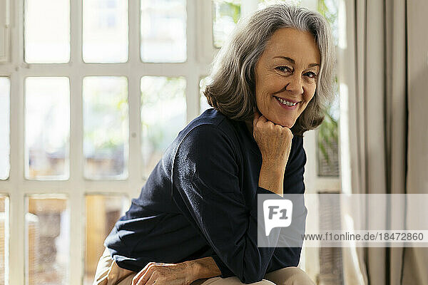 Happy mature woman sitting with hand on chin in front of window