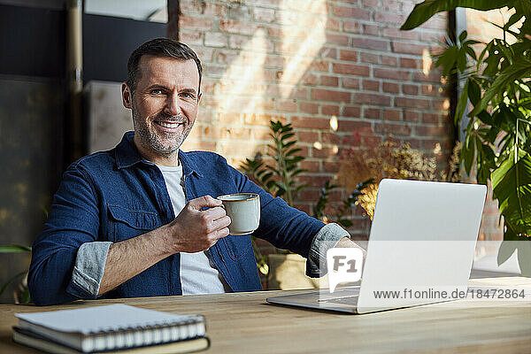 Smiling businessman with coffee cup sitting with laptop at loft office