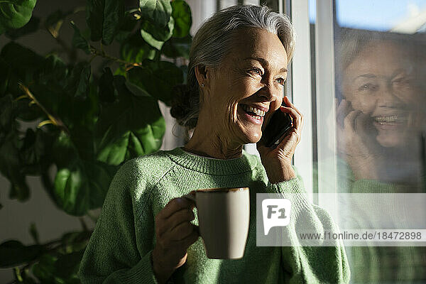 Happy mature woman talking on mobile phone by window at home