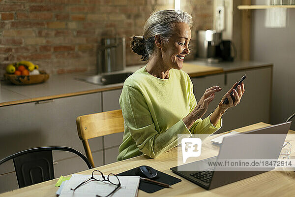 Happy mature freelancer using smart phone at desk