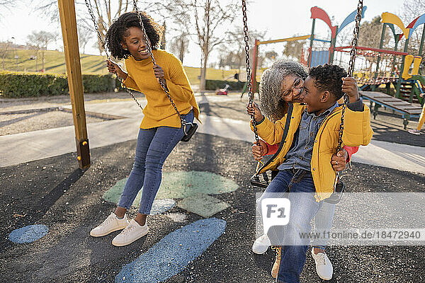 Glückliche Frau schaut Mutter zu  die ihren Enkel auf der Schaukel auf dem Spielplatz antreibt