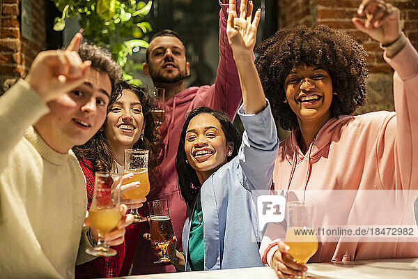 Happy multiracial friends with hands raised enjoying at restaurant