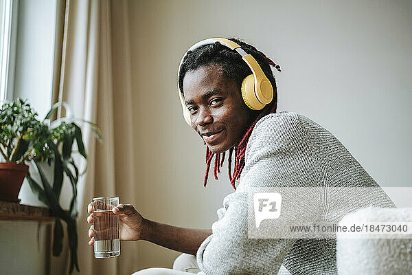 Smiling young man wearing headphones holding glass of water at home