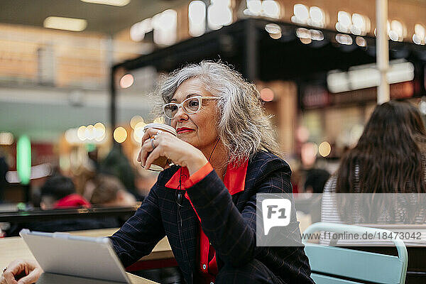 Nachdenkliche reife Geschäftsfrau sitzt mit Einwegkaffeetasse im Café