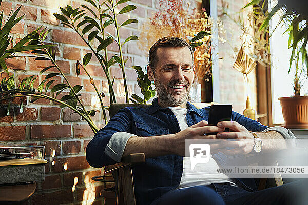 Happy man using mobile phone sitting on chair at home
