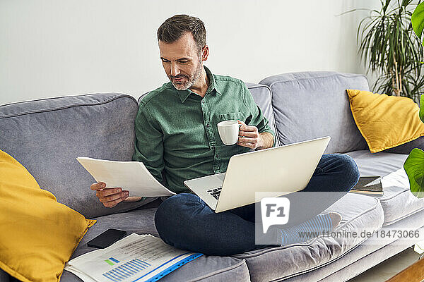 Man working from home looking at documents sitting cross-legged with laptop on sofa
