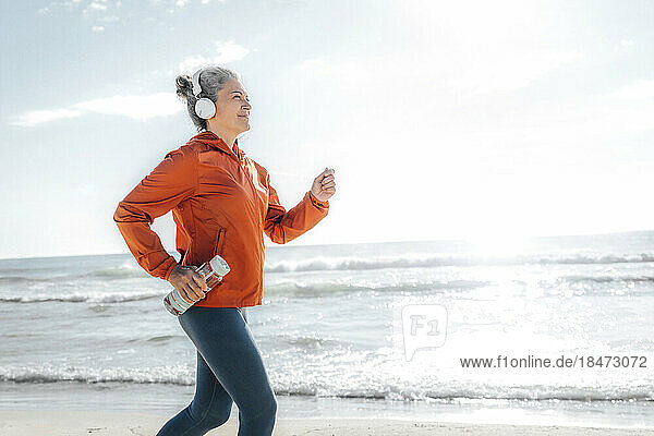 Smiling mature woman with water bottle jogging at beach on sunny day