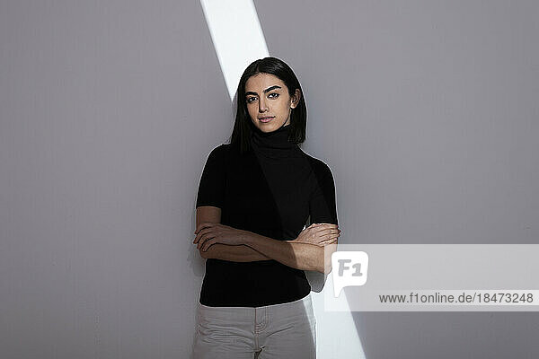 Young woman with arms crossed and light over face standing in front of white wall
