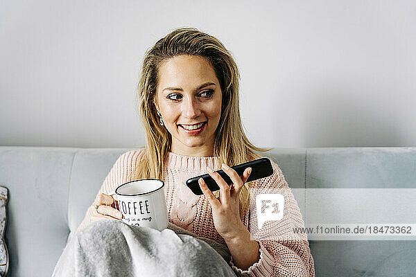 Happy woman with smart phone having coffee cup on sofa at home