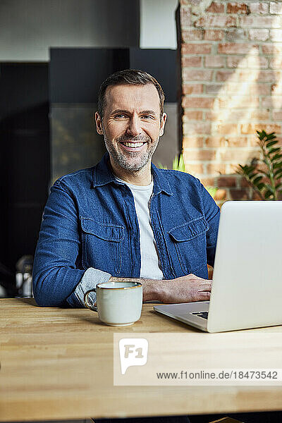 Smiling freelancer with laptop and coffee cup at desk in loft office
